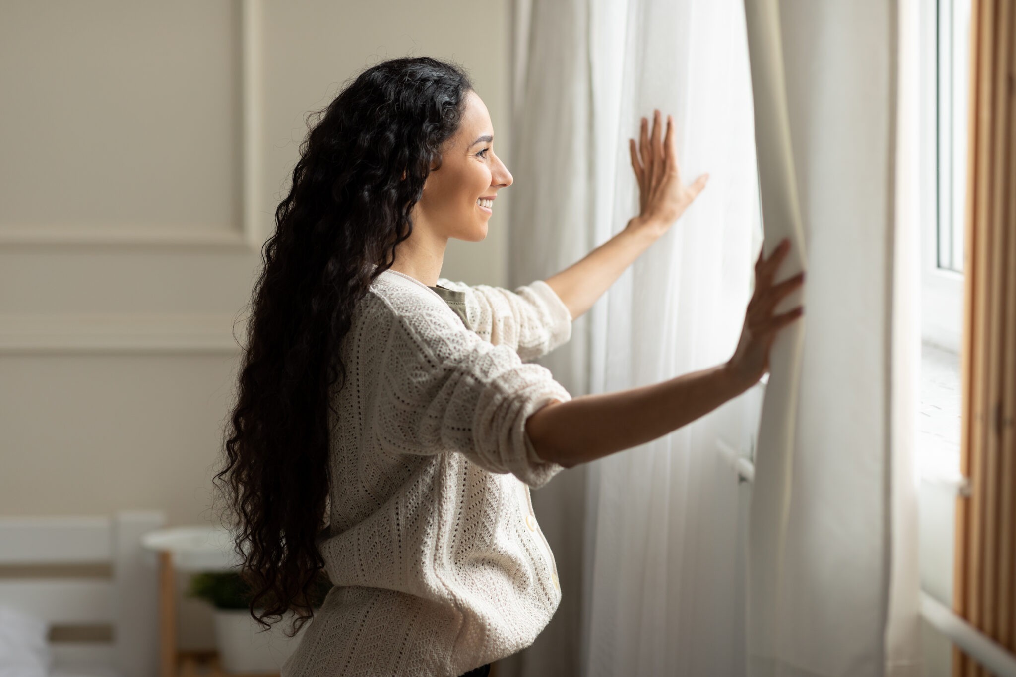 Smiling woman opening window curtains at home
